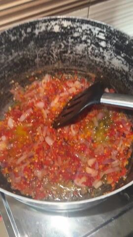 Here's a description of the video:

The video shows ingredients and cooking tools in a kitchen setting. First, a pot with red and white chopped vegetables (likely chili and onion) simmering in oil is shown. A black spatula is in the pot. The camera then moves to a dark, patterned countertop. On a white cutting board with a red border, there is a pile of chopped tomatoes. A large black cleaver rests on top of the tomatoes. Nearby are a red blender, a small bag of salt, and a bag of whole tomatoes.