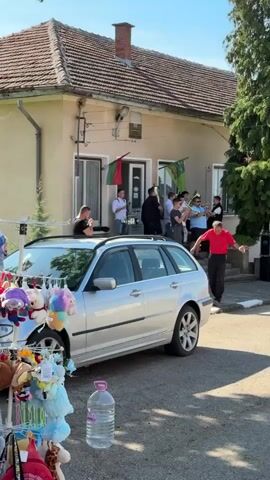 The video captures a lively street scene in front of a one-story building with a tiled roof. Several men are gathered on the steps and in front of the building’s doorway, playing brass instruments. They appear to be part of a brass band, dressed in various shirts and vests. Two Bulgarian flags are displayed on either side of the door. In the foreground, a silver station wagon with a sunroof is parked. A display stand laden with plush toys is set up next to the car, seemingly for sale. A man in a red shirt walks across the foreground, observing the band. Sunlight illuminates the scene, highlighting the colors and textures of the objects and people.