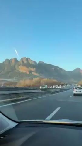 The video shows a view from the dashboard of a car driving on a highway. The highway has multiple lanes and there are other vehicles on the road, including cars, trucks, and a white van. A mountain range is visible in the distance against a clear blue sky. There's a trail of what seems to be a plane's exhaust in the sky.