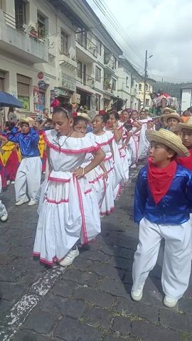 A group of children are dancing in a parade on a street with buildings on both sides. The children are dressed in traditional costumes. The girls wear white dresses with red trim, and the boys wear blue jackets, white pants, and straw hats. Some of the boys wear red neckerchiefs. The children are dancing in unison, moving their arms and legs in a coordinated manner. Some of the children are blowing whistles. Spectators line the street, watching the parade. The street is paved with cobblestones. A Coca-Cola sign is visible on one of the buildings.