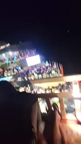 The video captures an outdoor evening gathering with a large screen displaying content. A crowd of people is seated, facing the screen, suggesting a viewing event. Flags are visible, possibly indicating a national or cultural event. The setting appears to be a venue with tiered seating and ambient lighting. The foreground shows a person in silhouette, relaxing and observing the scene.