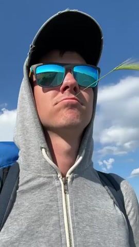 The video begins with a close-up, low-angle shot of a person's face against a bright blue sky with scattered white clouds. The person is wearing a grey hooded sweatshirt zipped up, a black baseball cap, and dark sunglasses with blue mirrored lenses. A piece of green grass is stuck to the person's upper lip. The person's expression is neutral. The camera then shifts slightly, showing more of the person's backpack, which is dark with blue accents. The background remains the sky and clouds, with a hint of a distant landscape. As the video progresses, the person's facial expression changes to a slight smile. The grass remains in place. The footage is handheld and appears to be a selfie. The lighting is bright and natural, suggesting daytime outdoors.