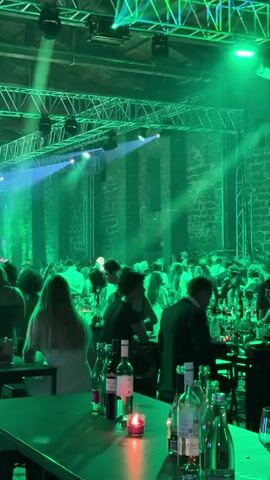 The video shows an indoor event with a large gathering of people seated at tables in a dimly lit space illuminated by green and white spotlights. The venue appears to be a large hall with exposed brick walls and a high ceiling with exposed truss systems and lighting equipment. Several tables are visible in the foreground and middle ground, set with bottles of wine and water, glasses, and small lit candles. People are seated at these tables, facing away from the camera for the most part, and some are engaged in conversation or eating. In the background, a stage area is visible with a person standing and speaking or performing, illuminated by spotlights. There are also other people standing and mingling in the background. The overall atmosphere appears to be that of a formal event, such as a banquet, wedding reception, or corporate function. The lighting and the presence of multiple tables suggest a seated dinner or awards ceremony.