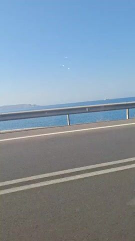 The video shows a view from a moving car. The camera is focused on the scenery to the right of the car. The road is asphalt, with a double white line. A gray metal safety railing is visible on the side of the road, separating it from the ocean. The ocean is a clear blue and extends to the horizon. An island or peninsula is visible in the distance on the left. A white ship is also visible in the distance, moving across the water. The sky is cloudless and a bright blue. There is no sound other than music. The car's side mirror is visible in the corner of the frame.