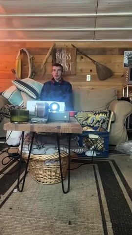 A young man is sitting on a couch in a room with a wood-paneled wall, watching a projector. The projector is set on a small, rustic table in front of the couch, along with a lit candle, a bowl, an Xbox, and an Xbox controller. The man has a headset around his neck. The wall behind the couch is decorated with items such as a life vest, a sign that reads "Bless This Nest," and an old broom. The room appears to be a living space or den with personal belongings scattered around. The camera slowly zooms out during the short video.