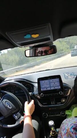 The video shows the interior of a car from a passenger's perspective. The driver, whose hands are visible on the steering wheel, is a person with sunglasses. The dashboard features a Ford logo and a touchscreen infotainment system. Two heart-shaped decals, one blue and one yellow, are affixed to the overhead console. The car is driving on a road with vegetation on the sides. Several other cars are visible on the road ahead. The light suggests it is daytime.
