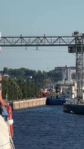 The video shows a canal with a ship moving through a lock. The ship is blue and white, with Russian text on its side that reads "ВОЛГО-ДОН". There is a crane structure over the canal, and buildings in the background. Several birds are flying in the sky and on the water. The water is dark blue and choppy. The sky is clear and blue. The overall scene appears to be a busy port or industrial area.
