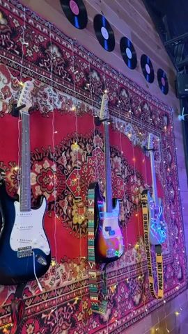 The video displays a wall adorned with a large, red, patterned rug. Four guitars are mounted on the wall, two electric and two bass guitars. They hang vertically, with their necks pointing upwards. There are string lights draped across the rug and guitars, and several vinyl records are displayed horizontally along the top of the wall. The lighting in the scene is dim, with purple and yellow hues present.