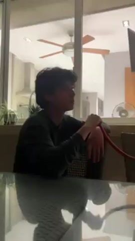 A young man with dark, wavy hair is seated at a glass table, inhaling from a hookah hose. He is wearing a dark long-sleeved shirt and is facing right in the frame. Behind him is a large window showing a room with a ceiling fan, kitchen appliances, and potted plants. The hookah hose is red. The table has a checkered pattern.