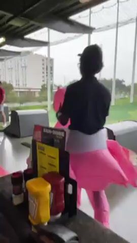 The video shows a person wearing an inflatable pink flamingo costume inside a Topgolf facility. The person is standing near a table with condiments and menus on it. In the background, other people are visible, and there is a view of a golf course and buildings. The lighting is indoor, and the video seems to be taken from a relatively close distance.