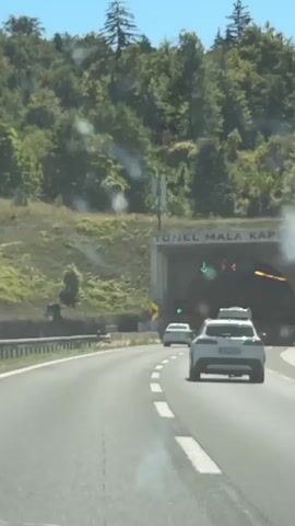 A car approaches a tunnel entrance marked "TUNEL MALA KAPELA". There are traffic lights above the entrance. One light shows a green arrow pointing down, indicating traffic can proceed. Other lights show red circles, likely indicating closures or restrictions in other lanes. The road ahead curves slightly to the right as it enters the tunnel. Several other vehicles are visible ahead of the car, including a white SUV. The tunnel is flanked by a green, tree-covered hillside. The sky is clear and blue.