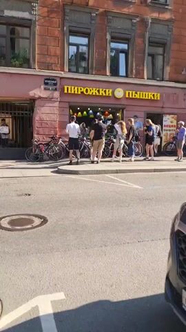 The video shows a street scene with a building facade that has a sign reading "ПИРОЖКИ ПЫШКИ" in large yellow letters. Below the sign, there is an entrance with several people standing in line, and bicycles are parked to the left of the entrance. The people in line appear to be customers. The building is made of red brick and has windows on the upper floors. The street in front of the building has a manhole cover and a portion of a car is visible in the foreground. The weather appears to be sunny.