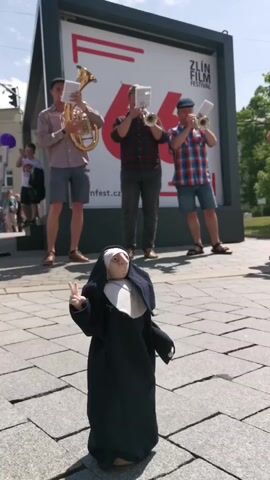 The video shows a group of musicians playing in front of a Zlin Film Festival poster on a sunny day. The musicians consist of three men, one playing a tuba, and two playing trumpets. In the foreground, a doll dressed as a nun stands with its hand raised in a peace sign. The scene is set outdoors on a stone-tiled walkway. Other pedestrians are visible in the background.
