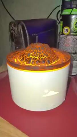 A white, cylindrical humidifier resembling a volcano is positioned on a burgundy surface. The humidifier has an orange glowing top that is textured to resemble lava. Steam or vapor emanates from the top of the humidifier. The base of the humidifier has an illuminated orange symbol that resembles a cloud with a sun above it. Behind the humidifier is a black bag, a blue object, and a can with a warning label that says "18" inside of a yellow circle. The surface appears to be slightly reflective.