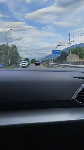 The video captures a first-person perspective from inside a car driving on a highway. The road is flanked by lush green trees and mountainous terrain, with a clear blue sky dotted with fluffy white clouds. A dark-colored sedan is visible ahead on the road, and a white SUV is passing by on the left lane.

A prominent blue sign with white text is visible on the right side of the road. The text reads "controllo elettronico della velocita," which translates to "electronic speed control" in English. This indicates the presence of a speed camera or other electronic speed monitoring system. Another smaller sign with the number "70" is also visible, likely indicating a speed limit.

The overall setting suggests a daytime drive in a scenic, rural area, possibly in the Valtellina region of Italy given the location context. The weather appears pleasant, with good visibility. The movement of the car creates a dynamic view of the landscape. No people are clearly discernible within the frame, and the focus is on the road, the surrounding environment, and the car's dashboard. The atmosphere is one of typical highway travel through a beautiful natural setting.