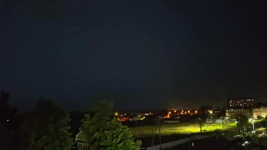 This video shows a night scene of a town with buildings and trees under a dark sky, likely during a thunderstorm, as evidenced by occasional flashes of lightning illuminating the sky and landscape.