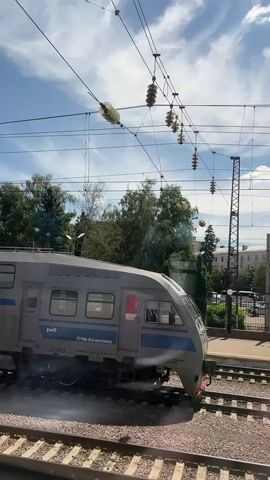 This media captures a train at a station on a sunny day with a bright blue sky and scattered clouds. The train, a modern, grey passenger car with a blue stripe, is stationary or moving slowly on train tracks. The front of the train has the designation "ПЧМ-КОЧЕТОВКА" and "АС-01 №35" written on it. There is steam or smoke emitting from the front underside of the train.

In the background, large, multi-story buildings with white and yellow facades and ornate architectural details suggest an urban or town setting. Lush green trees are visible behind the buildings, and overhead, complex electrical wires for the train system crisscross the sky, supported by insulators. The platform of the train station is also visible. The overall impression is of a typical day at a Russian railway station.