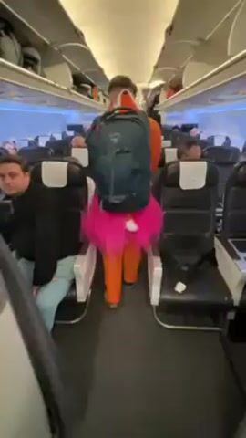Inside an airplane, a man walks down the aisle toward the front of the plane. He wears an orange jumpsuit, a pink tutu, and a large gray backpack. Passengers are seated in the rows of seats on either side of the aisle. Most are facing forward. Many are in dark clothing. The cabin's overhead storage is visible. The plane appears to be mostly full.