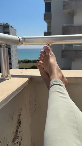 A person wearing light green leggings rests their bare feet on a concrete balcony railing, toes pointed upwards, with pink nail polish on each toe. In the background, there is a clear blue sky, buildings, and a glimpse of the sea. A metal railing runs horizontally along the balcony. The scene is brightly lit by sunlight.