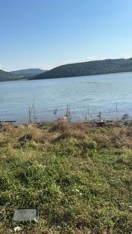 The video captures a serene daytime scene at a lake in Sava, Bulgaria. The sky is a clear, bright blue, indicating fair weather. The lake's surface is calm, with gentle ripples reflecting the sunlight. Rolling, tree-covered hills form the backdrop, suggesting a rural or natural environment.

In the foreground, the grassy shore is dry and somewhat overgrown, with patches of taller, withered vegetation. A fishing rod is propped up on the bank, indicating someone is likely fishing. A dark, flat object, possibly a tackle box or equipment, rests on the ground near the grass. A bright red object, possibly a piece of litter or equipment, is visible near the water's edge.

The primary activity observed is a person swimming in the lake. Their head is visible above the water, and they appear to be some distance from the shore. The presence of the swimmer adds a dynamic element to the otherwise still landscape. There is no visible text in the video. The overall mood is peaceful and relaxed, capturing a moment of outdoor recreation on a pleasant day.