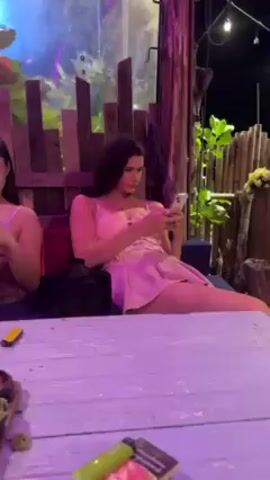 Two young women sit side-by-side at a white wooden table, each absorbed in their smartphones. Both women wear pink dresses with thin straps. The woman on the right has curly dark hair, while the woman on the left has straight dark hair. On the table are a lighter, a pack of cigarettes, and a small dish with what appears to be some plant matter. The background features wooden structures and greenery, suggesting an outdoor or semi-outdoor setting, possibly a bar or restaurant. The scene is lit with pink and purple lights, creating a relaxed and casual atmosphere.