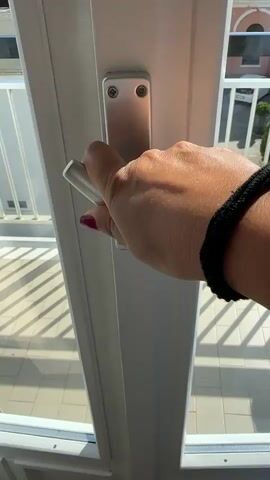 The video begins with a close-up of a person's hand turning a silver handle on a white door or window frame. The camera then pulls back to reveal a view from a balcony. The balcony has white railings and is tiled with beige squares. Beyond the balcony, there is a paved area with potted plants, a white car, and a building with pink and white facades. In the distance, a line of tall green trees is visible against a blue sky, with the sea and a beach behind them. The sun is shining, casting shadows on the balcony tiles.