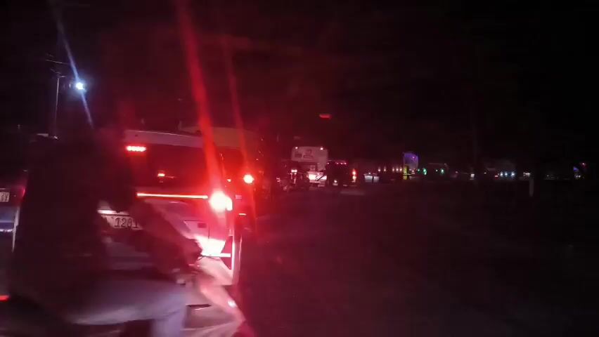 The video shows a nighttime scene of traffic on a road. The camera is likely mounted on a motorcycle, positioned behind a white SUV with the license plate "BL 1281 NI." The motorcycle rider is visible in the lower center of the frame, wearing a dark shirt and jeans. Several vehicles, including trucks and cars, are visible further down the road, their lights creating a line of illumination. Streetlights are also present along the roadside. The general atmosphere suggests a congested traffic situation at night.
