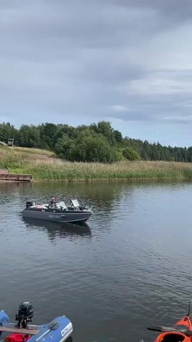 A gray motorboat with a single person at the helm moves across calm, dark water. The boat has a silver engine mounted at the stern and a windshield at the front. The person is wearing a dark jacket and a hat. The boat has Russian registration numbers visible on its side. In the background, a lush green shoreline is lined with tall, dry reeds and dense trees. The sky above is partly cloudy with a light gray hue. In the foreground, the bows of two other boats are visible, one is a yellow kayak and the other is a gray inflatable boat with an outboard motor.