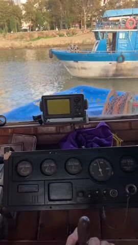 The video shows a first-person view from inside a boat's cabin, looking out through the windshield. In the foreground, a person's hands are on a wooden steering wheel. The dashboard has several analog gauges, a screen displaying what appears to be a fish finder or GPS, and a purple cloth. Outside, a blue and white boat is passing by on a body of water. The background shows a tree-lined shore with buildings and a flag. The video is set in daylight.