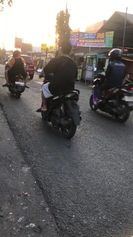The media file captures a street scene in Balonggabus, Indonesia, likely during the late afternoon or early evening due to the warm, low sunlight. The setting appears to be a moderately busy road, with asphalt surfacing and a paved shoulder. Several individuals are riding motorcycles, a common mode of transportation in the area.

The subjects are primarily people on motorcycles, seen from behind. In the foreground, a motorcycle with a rider and passenger is moving away from the camera. The passenger's clothing is blurred due to motion. Further down the road, other motorcycles are also visible, with riders of varying ages and genders. One rider wears a helmet. The motorcycles themselves are standard models, with some appearing to be scooters.

The activity is the general movement of traffic on a road. The scene suggests a daily commute or local travel. The weather seems fair, with clear skies allowing for the bright, directional sunlight.

Notable details include the distinct appearance of the road, with its textured asphalt. In the background, there are buildings and stalls lining the street, indicative of a commercial or residential area. A prominent banner hangs above one of the stalls, featuring text in Indonesian. The overall impression is one of everyday life in a Southeast Asian locale.

Visible text includes "SPB", "SUSU", and what appears to be a date range "2020-2027" on the banner in the background. The word "SUSU" means "milk" in Indonesian. "SPB" could be an abbreviation for a company or organization. The context of the banner and the surrounding stalls suggests it might be related to local businesses or public services. There is no discernible emotional tone conveyed, as the focus is on the activity and environment.