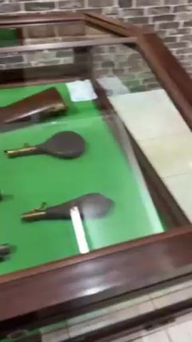 The video shows a glass display case with various old firearms and related items on a green surface. There are several long guns, possibly muskets or rifles, arranged in rows.  Next to some of the firearms are small identification labels. In addition to the long guns, there are also powder flasks. The environment appears to be a museum or historical exhibit, with a brick wall visible in the background.