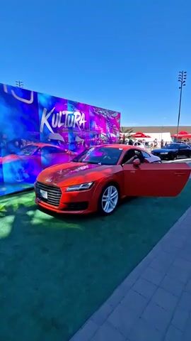 A red Audi TT is parked on a green carpet. The driver's side door is open, and a person is partially visible inside the car. Behind the car is a large blue and purple banner with the word "KULTURA" written in large white letters. There are other cars and people in the background, suggesting an outdoor event. The sky is clear and blue.