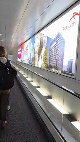 A woman wearing a backpack and khaki pants walks on a moving walkway alongside a large illuminated advertisement. Another person is visible in the distance on the same walkway. The walkway is enclosed by glass on one side, offering a view of an outdoor area with vehicles. The ceiling is made of metallic panels with embedded lighting.