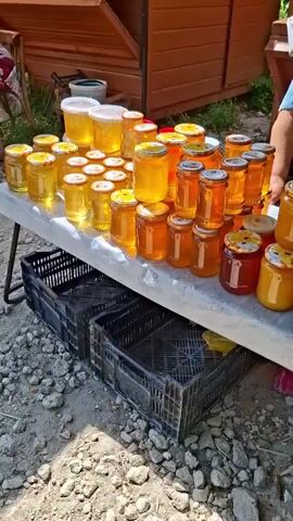 The video shows an outdoor market stall selling jars of honey and other preserved goods. A woman stands behind the stall, wearing a blue towel around her neck and a black and white polka dot shirt. She is smiling and looking at the camera. The stall is set up with multiple tables displaying rows of glass jars filled with golden-colored honey, as well as jars of red preserves. There are also baskets containing dried herbs and other packaged goods. In front of the stall, there are black crates and a red cooler. The ground is covered with gravel and stones. The background shows a wooden structure, likely part of the market stall or a nearby building. The overall atmosphere appears to be that of a local market or roadside stand.