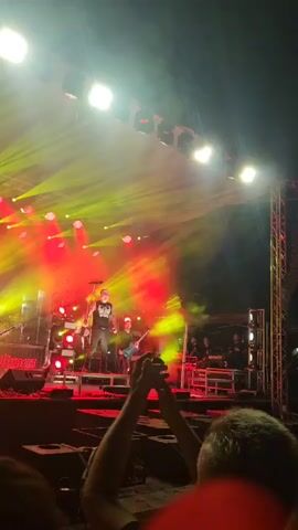 A concert is taking place on a stage. A male singer is on stage, wearing black clothing and holding a microphone. He is standing on a raised platform, leaning forward. There are red and yellow lights illuminating the stage, with smoke effects. Other band members are also on stage, playing instruments, including guitars. The audience is visible in the foreground, with some people holding up their phones.
