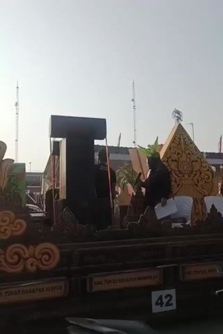 A float is decorated with ornate designs and a large black structure, possibly a speaker system, in the center. Three individuals are visible on the float. One person, wearing a green headdress, is speaking into a microphone, holding a piece of paper. Another person stands behind the speaker system, and a third person is partially visible on the left side of the float. The background shows a bright sky with the sun, and some distant structures and antennas. A sign with the number 42 is visible at the bottom of the frame. The overall setting appears to be an outdoor event or parade.