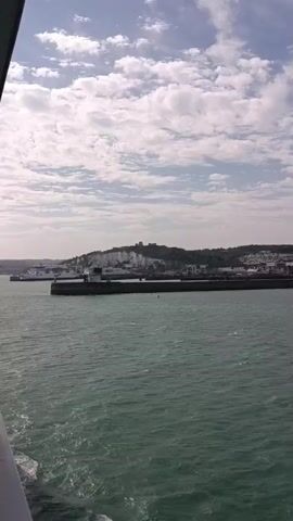 The video shows a choppy, green sea in the foreground. In the middle ground, there is a harbor with a breakwater, a ferry terminal, and buildings on the shore. In the background, there are white cliffs, some with buildings and structures on top, and a castle-like structure on a hill. The sky is partly cloudy with white clouds against a blue sky. The video appears to be taken from a moving vessel, likely a ferry, as it approaches or departs the harbor. The lighting suggests it is daytime, possibly late morning or afternoon, with the sun behind the camera. There are no people or clear actions visible in the footage.