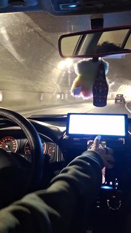 The video is shot from inside a car driving through a tunnel. The dashboard is visible, including the steering wheel, speedometer, a large screen, and a phone mounted on the center console. The phone displays a music playlist. A fuzzy rainbow-colored decoration and an air freshener hang from the rearview mirror. The tunnel walls are light gray and illuminated by artificial lights. Another car is visible in the distance, moving in the same direction. A hand is visible on the steering wheel. The audio includes music.