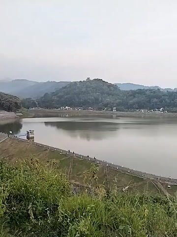The video presents a wide, panning shot of a large, tranquil reservoir or lake. The camera moves smoothly from left to right, revealing the expansive body of water and its surrounding landscape.

In the immediate foreground, lush green grass and foliage are visible. A significant dam wall or embankment curves along the edge of the reservoir, and several people are seen casually walking along its top, enjoying the view. A small, multi-story concrete building, possibly a control tower or pump house, stands prominently on the dam.

The water of the reservoir is calm and reflective, mirroring the soft, pale sky. In the midground and background, undulating hills covered in dense green trees frame the reservoir. Some scattered buildings are visible nestled among the trees on the far side of the water.

The sky is overcast and hazy, suggesting either early morning or late afternoon with diffused lighting. The overall scene conveys a sense of peace and natural beauty in a hydrological setting. The location, Jambeyan, Indonesia, is consistent with the verdant, hilly terrain. No visible text or distinct events are noted, only the serene landscape and leisurely activity of a few individuals.