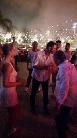 A group of people are dancing outdoors at night. String lights are visible in the background. Some people are wearing white shirts. There is smoke or fog in the air. One woman in the foreground is wearing a white dress and appears to be dancing with a man in a white shirt. Other people are dancing or standing around.
