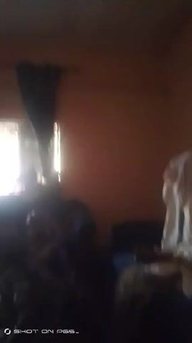 The video shows a poorly lit room with several people gathered inside. The people appear to be seated, and some are wearing patterned clothing. A window is visible in the background, letting in a bright light that creates a strong contrast with the rest of the room. The overall scene is dark and blurry, making it difficult to discern specific details. The video appears to be taken from a low angle, moving slightly.
