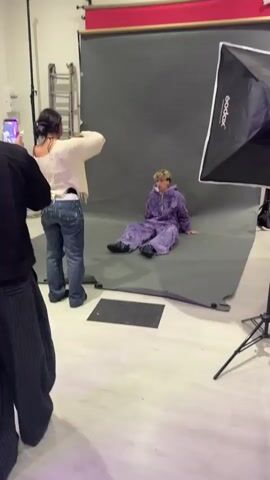 A young adult male is sitting on a grey backdrop in a photo studio. He is wearing a purple hooded jumpsuit with a ragged design. He has light blonde hair and is looking towards the camera. Two people are positioned in front of him, seemingly taking photos with their phones. One person is partially obscured, wearing a white top and jeans. The other person is wearing a black jacket and striped trousers. A large Godox softbox is on the right side of the frame. A ladder and a metal rack are visible in the background. The studio has a white floor.