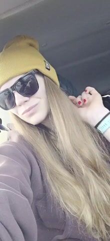 Here's a description of the video:

A woman is in a car, likely the driver. She's wearing a mustard-yellow beanie, sunglasses, and a brown hoodie. She has long, straight hair. At one point, she smiles. Music plays in the background. The interior of the car is visible. She wears a smartwatch on her left wrist. Her nails are painted red.