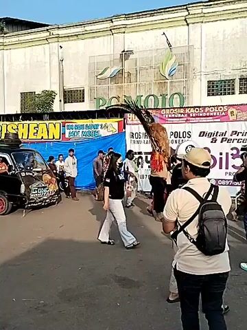 The video captures an outdoor market scene in Sragen, Indonesia, during the daytime under a clear blue sky. Numerous people, including adults and children, are seen walking casually through the area, browsing or passing by.

In the foreground and midground, a small black pickup truck is parked, facing towards the right side of the frame. Temporary market stalls with colorful tarpaulin roofs (blue and pink) line the street, indicating a bustling commercial activity. A large, light-colored building, possibly a factory or warehouse, stands prominently in the background, with palm trees visible in front of it.

Visible text on a large blue banner behind the truck reads "SHEKAYLA BISHEAN" or similar, with a sub-line stating "100 RIBU DAPAT 4 KAOS" (100 thousand [Rupiah] gets 4 shirts) and "MIX MODEL," suggesting a clothing sale. Another banner further down the street displays "GROSIR KAOS" (Wholesale T-shirts). The atmosphere appears lively but unhurried, with people enjoying the market.