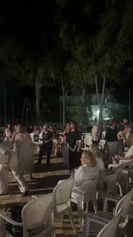 This is a vertical video filmed at night outdoors, likely in a courtyard or patio area with trees overhead. A group of people, mostly women, are dancing in the center of the frame. Most of the dancers are wearing white clothing, with a few wearing black. Some are dancing in pairs, while others are dancing individually or in small groups. Several people are seated in white plastic chairs around the perimeter of the dancing area, watching the dancers. The ground is paved with what appears to be interlocking stones. The overall atmosphere is festive and celebratory.