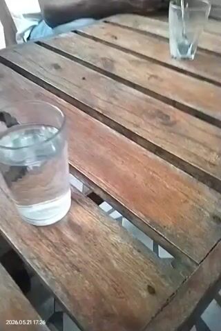 The video shows several men sitting at wooden tables in what appears to be a cafe or outdoor setting. The camera focuses on a table with glasses of water, cups of coffee, a smartphone, and a pack of cigarettes. One man is visible using a phone. Another man, wearing a white shirt and blue jeans, is sitting and talking. The camera pans around to show other men seated at different tables. The overall environment seems relaxed, with people enjoying drinks and conversation. The date and time "2026.05.21 17:26" are stamped in the lower left corner of the video.