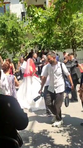 This is a video of a wedding celebration outdoors. A bride in a white wedding dress and red veil is surrounded by guests who are dancing and celebrating. Several men in suits are also present, as are other guests dressed in casual attire. The scene appears to be in a street or courtyard with trees and buildings in the background. The overall atmosphere is festive and joyful.
