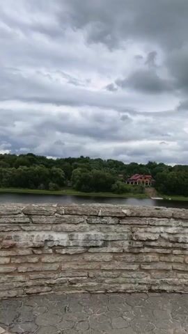 A vertical video shows a view over a stone wall, focusing on a river and a forested shoreline with a building visible on the opposite bank. The sky is cloudy. A young girl with long blonde hair stands with her back to the camera, looking out at the river. She is wearing a black t-shirt and white shorts. She raises her right hand and appears to be waving or gesturing.