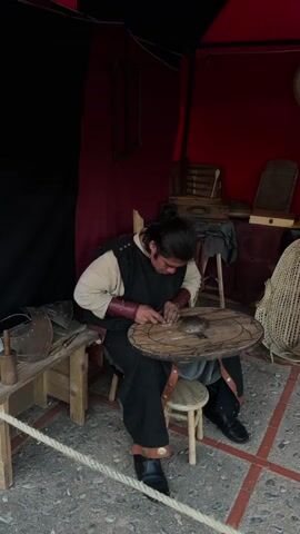The video shows a man in a medieval outfit sitting in a booth. He is working on a wooden shield. The man smiles at the camera, then continues working. In the background are tools, boxes, a wicker chair, and a woven basket. The booth is red and black. The floor is covered in square tiles.