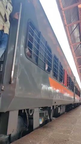 The video shows a train at a station. The train is gray and orange with black barred windows. People are visible inside the train looking out the windows. The train doors are red and some are open. The train tracks are visible below the train and the platform is made of textured metal. The train appears to be moving slowly or starting to move. A roof structure is visible above the platform.